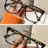 A pair of tortoiseshell reading glasses with clear lenses being held by a hand against an orange knitted background.