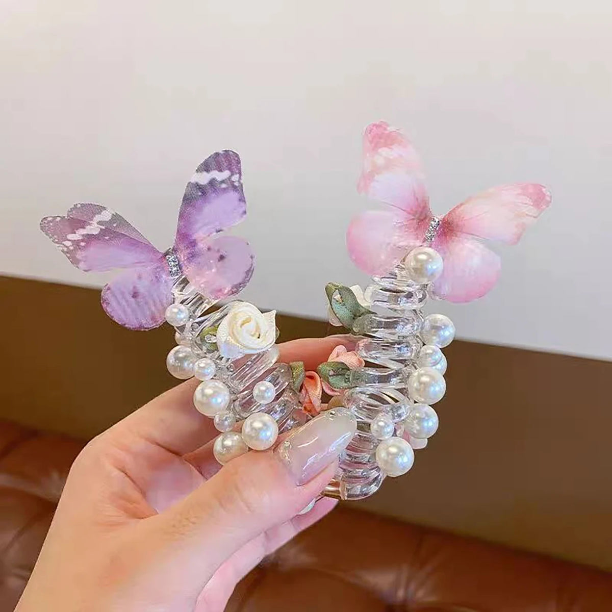 A hand holds a delicate hair rope featuring pink and purple fairy butterfly flowers and pearl beads.