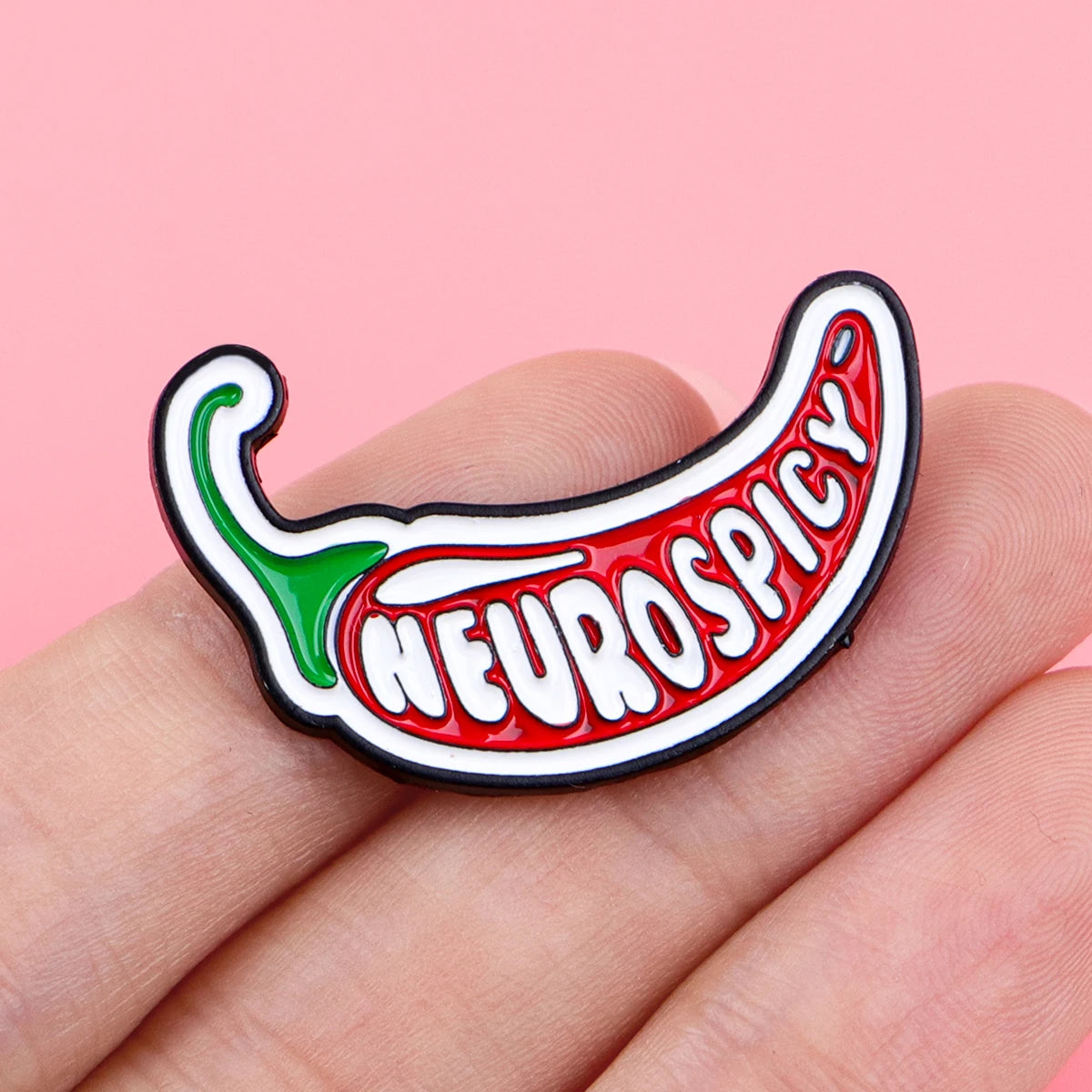 A red chili pepper shaped enamel pin with the word NEUROSPICY on it, held by a hand.