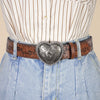 A close-up of a woman's torso showing a striped shirt, denim pants, and a heart-shaped retro embossed leather belt.