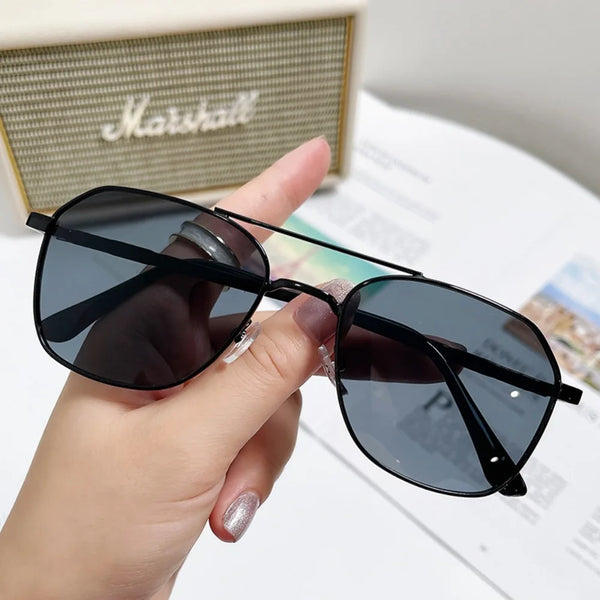 A hand holds a pair of black square sunglasses with a metal frame against a light background.
