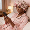 A woman wearing a pink collared shirt and a leopard print hair drying cap is looking at her phone in a bedroom setting.