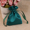 A teal satin drawstring pouch tied with a green ribbon rests next to a potted pink flower arrangement.