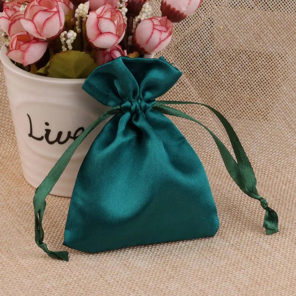 A teal satin drawstring pouch tied with a green ribbon rests next to a potted pink flower arrangement.