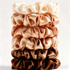 A stack of five satin hair scrunchies in various shades of nude, peach, and brown.
