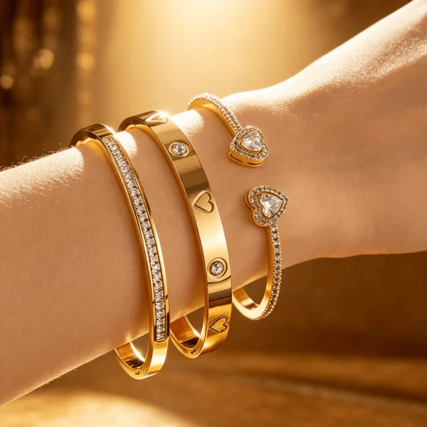 A hand wearing a set of three gold-toned bracelets, including a chain bracelet and two heart-shaped rings, is shown in soft l
