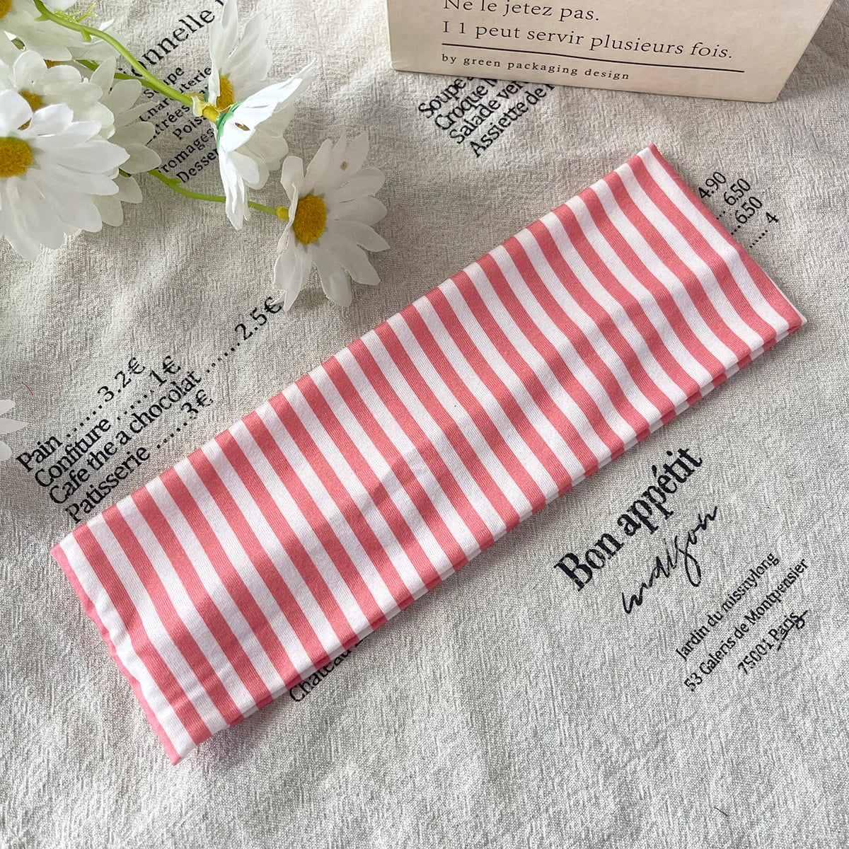 Un élastique pour cheveux rayé rose et blanc posé sur un tissu blanc avec des fleurs et du texte.