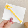A yellow hair elastic and rubber band remover tool is shown against a white card, held by a hand.