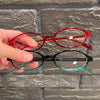 Une main tient une paire de lunettes de mode ovales rétro Y2K, avec des montures rouges et vertes.