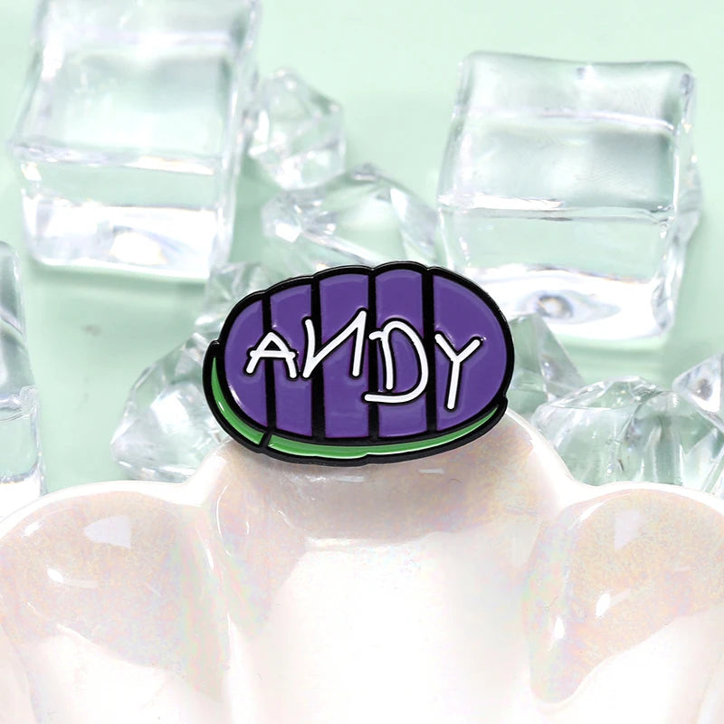 دبوس طية ياقة يحمل اسم ANDY بخط بنفسجي على خلفية بيضاوية داكنة