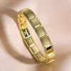 A close-up shot of a gold stainless steel charm bracelet on a light pink background.