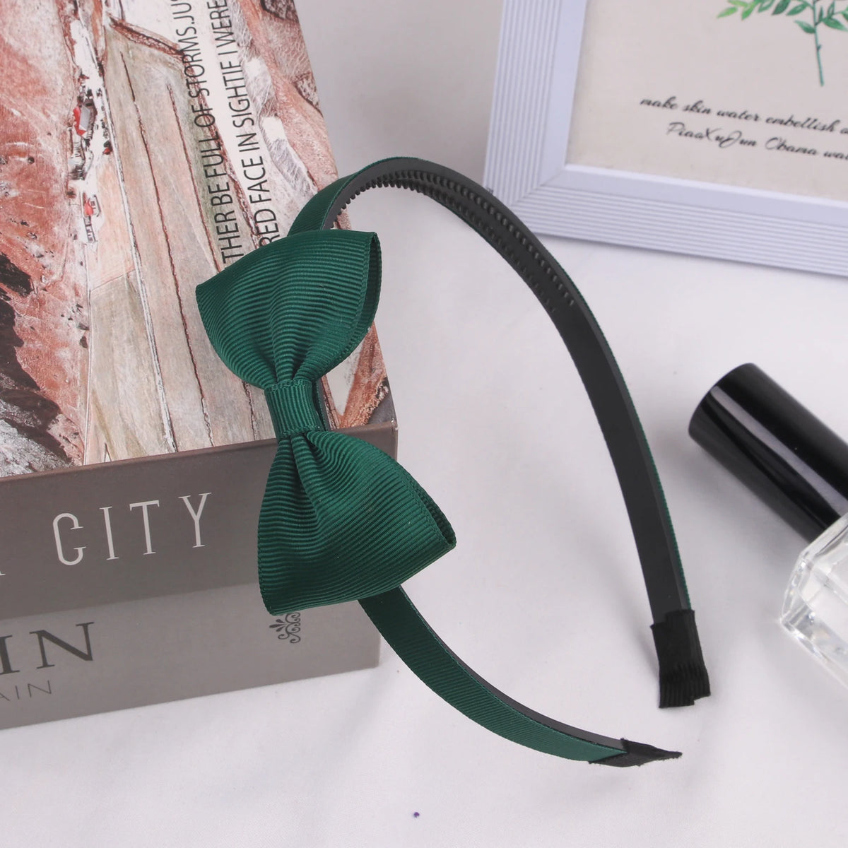 A dark green grosgrain ribbon hair bow is displayed next to a book and a small glass container.