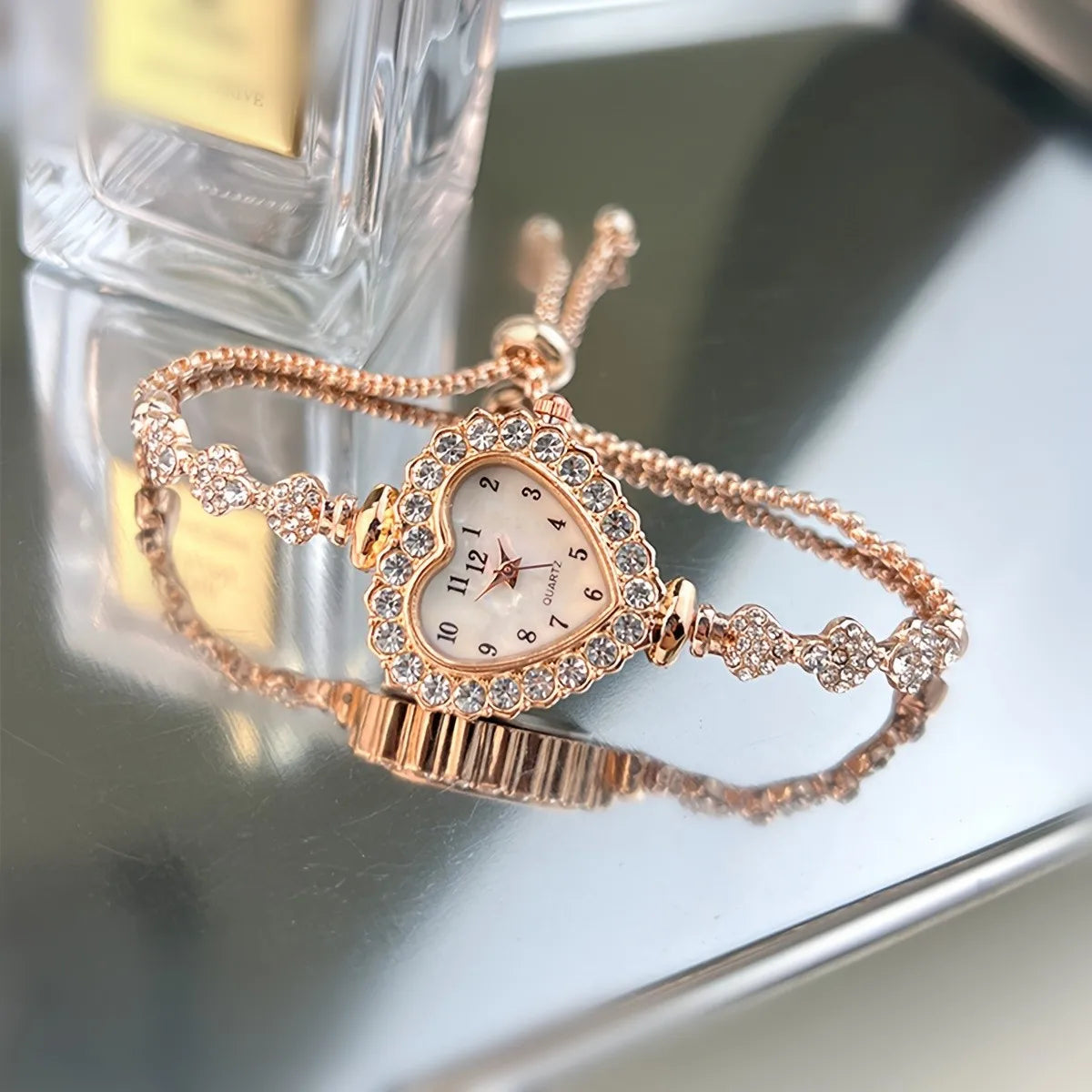 A close-up of a rose gold sparkling heart rhinestone quartz watch on a silver surface.