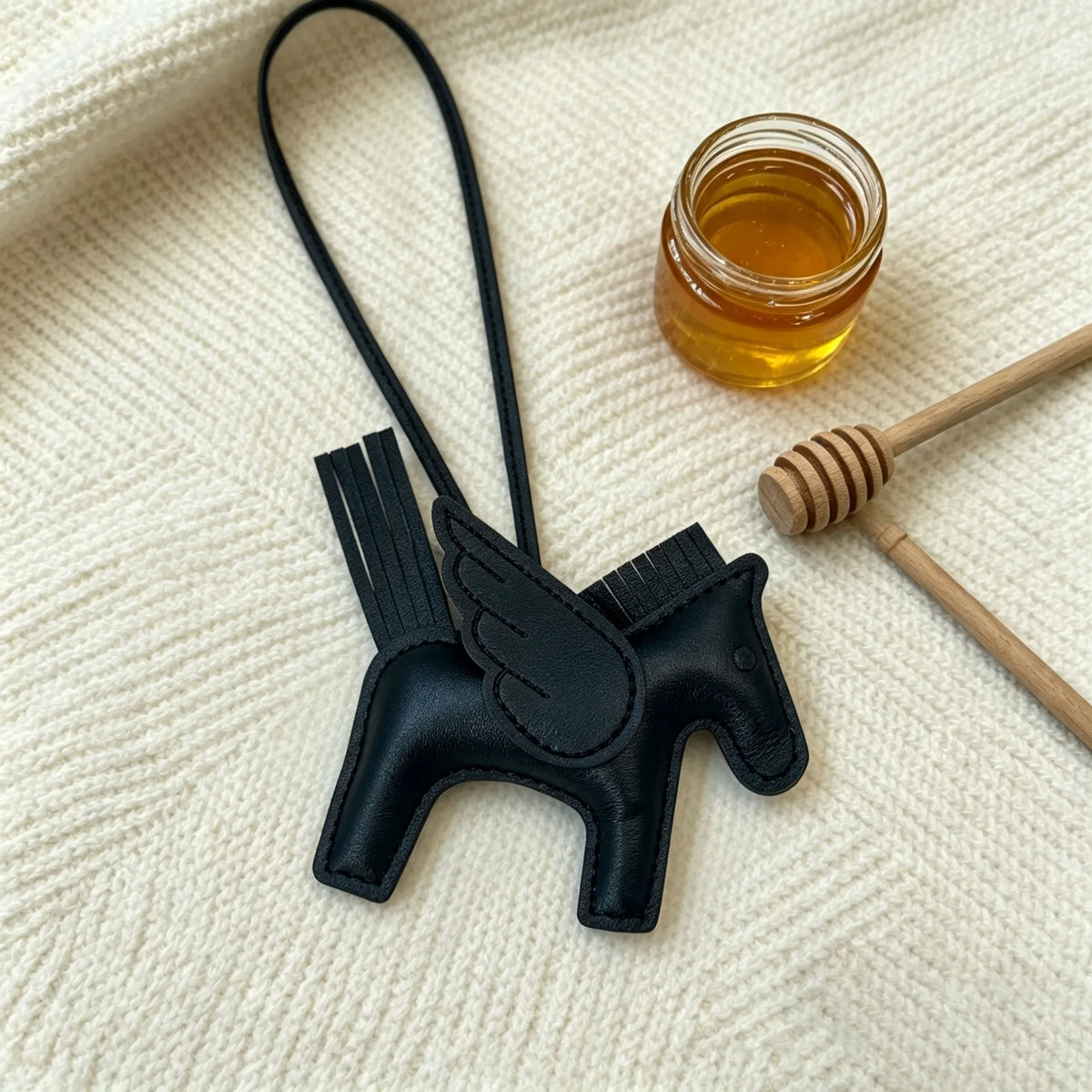 A black elegant horse pendant charm with a leather strap is displayed on a cream knitted fabric next to a jar of honey.