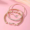 A set of three gold-toned heart charm bracelets with clear crystals displayed on a pink background.