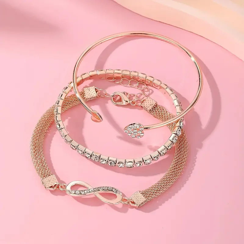 A set of three gold-toned heart charm bracelets with clear crystals displayed on a pink background.