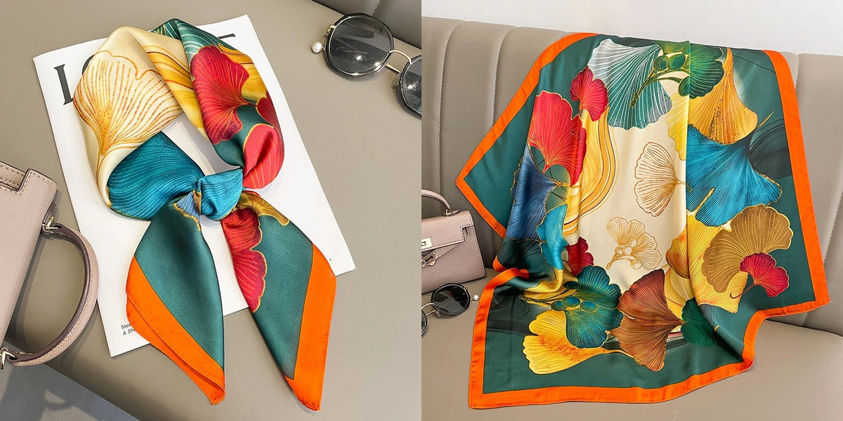 A vibrant floral print silk scarf featuring shades of teal, orange, yellow, and green draped on a light gray surface.