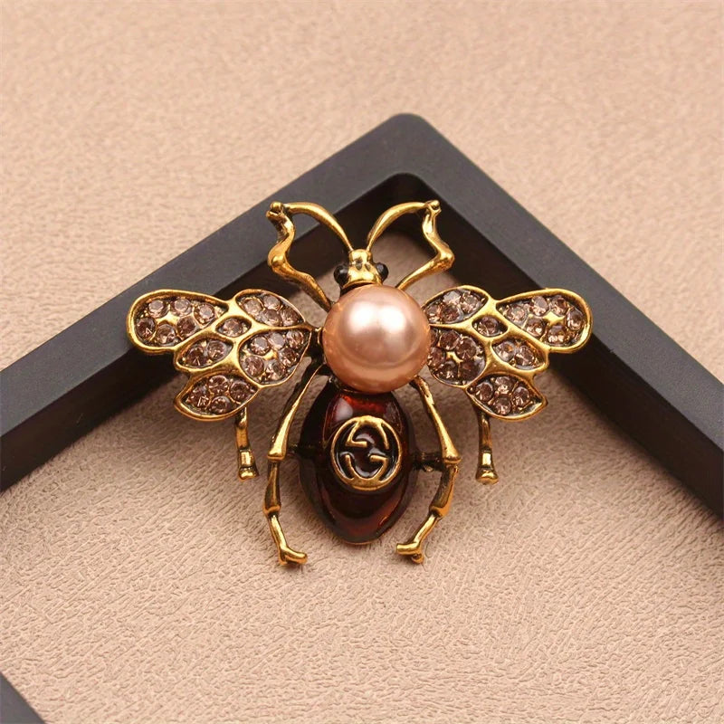 Broche couleur or avec motif d'abeille, perle rose et accents dorés sur fond noir.