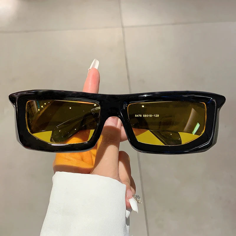 Una mano sostiene unas gafas de sol negras con lentes tintados en dorado y un aro metálico contra un fondo claro.