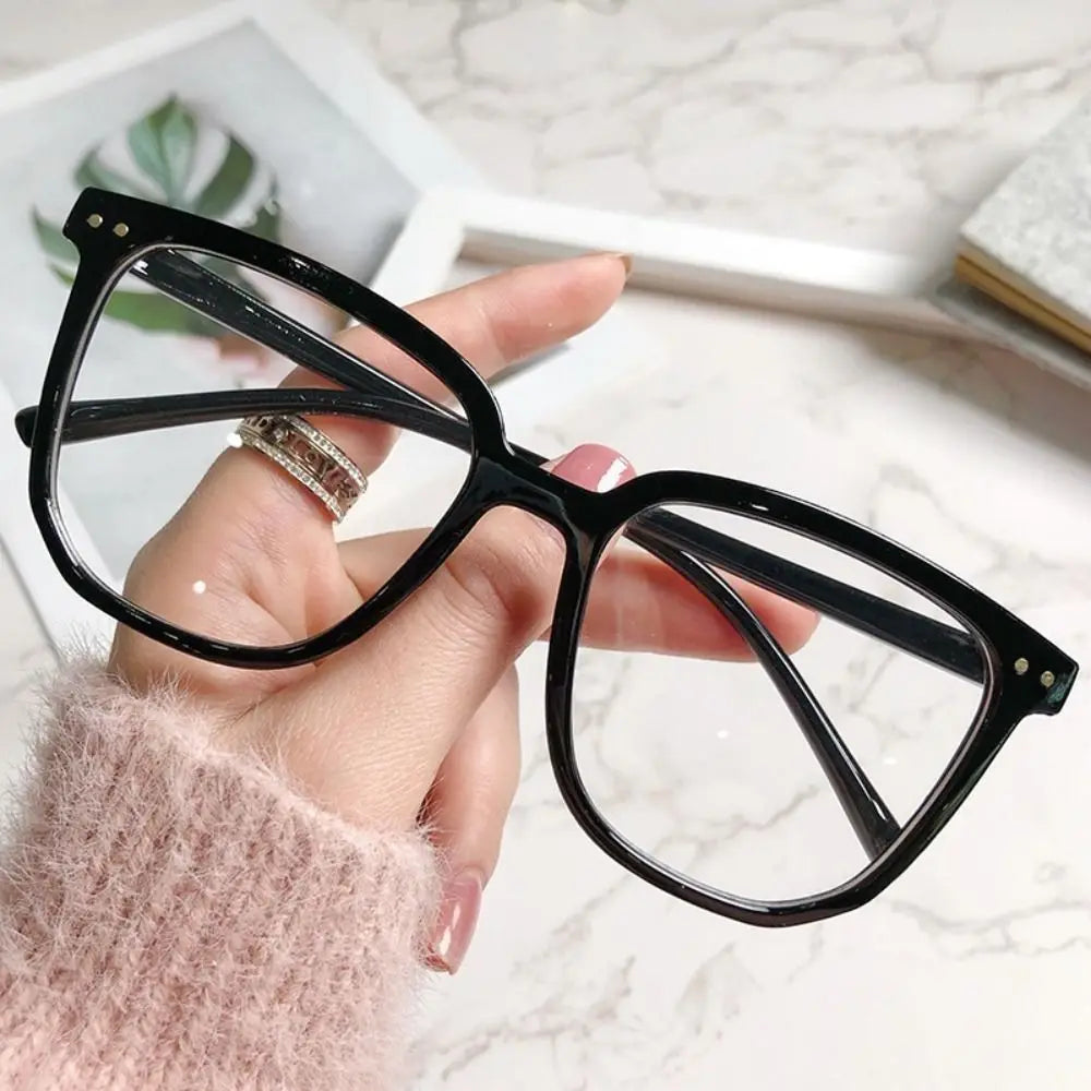 A hand holds a pair of black, large square anti-blue light spectacles against a white marble background.