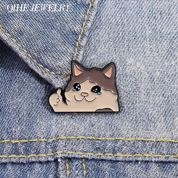 A small, cute cat enamel brooch pin with a smiling expression is pinned onto a denim fabric.
