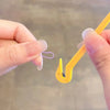 A hand is using a yellow tool to remove a purple elastic band from a fingernail.