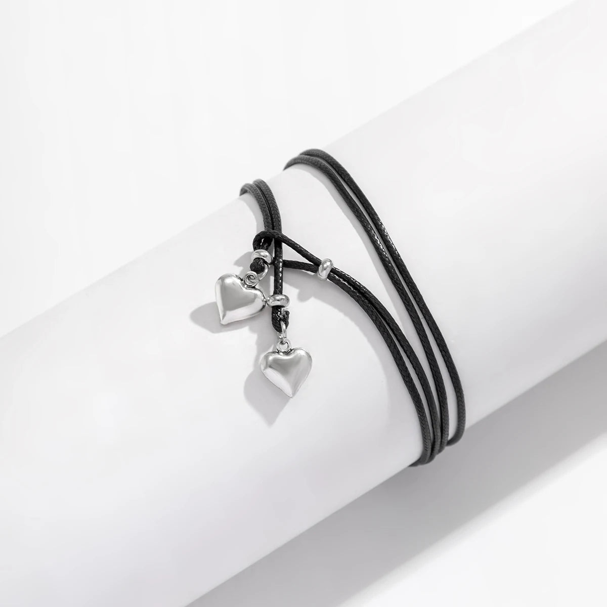 A black leather anklet with silver heart charms is shown on a white wrist.