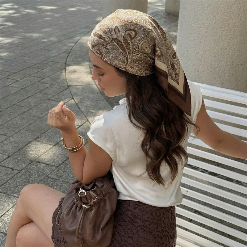 امرأة ترتدي وشاحاً من الساتان البيج بنقشة بيزلي وبلوزة بيضاء تجلس في الخارج.