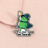 Un pin de esmalte con un mago rana verde con sombrero puntiagudo y vara sobre un fondo de tela rosa claro.