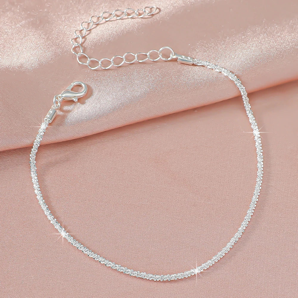 A delicate silver-plated chain anklet adorned with clear cubic zirconia stones, displayed on a pink fabric.