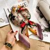 A luxury silk scarf in floral print with red and green accents is displayed on a white card next to a gold watch and beige fa