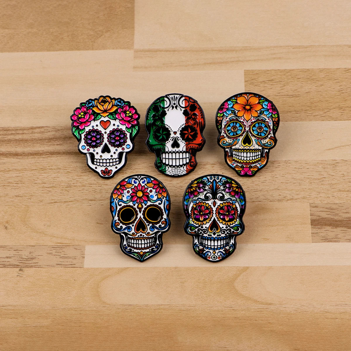 A collection of seven colorful Mexican skull enamel pins arranged on a light wooden surface.