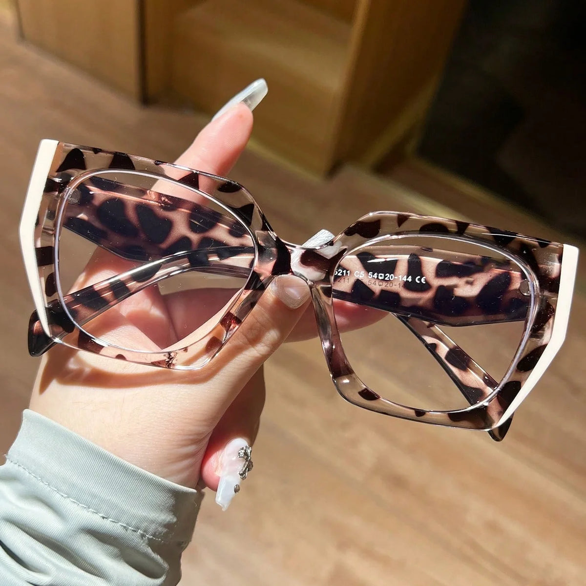 A hand is holding a pair of clear acetate glasses with a leopard print pattern.