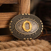 A vintage western belt buckle with an ornate gold design set in a dark metal frame, resting on a coarse brown textile.