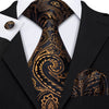 A black silk jacquard tie set featuring an ornate gold paisley pattern worn with a black suit jacket and white shirt.