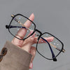 A hand holds a pair of black and purple anti-blue light metal eyeglasses against a gray background.