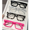 A display of five pairs of reading glasses in various styles and colors including black, clear, tortoiseshell, and pink.