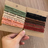 Eine Sammlung von Boho-Elasthebändern in verschiedenen Grüntönen, Beige und Pink auf einem Kartonanhänger.