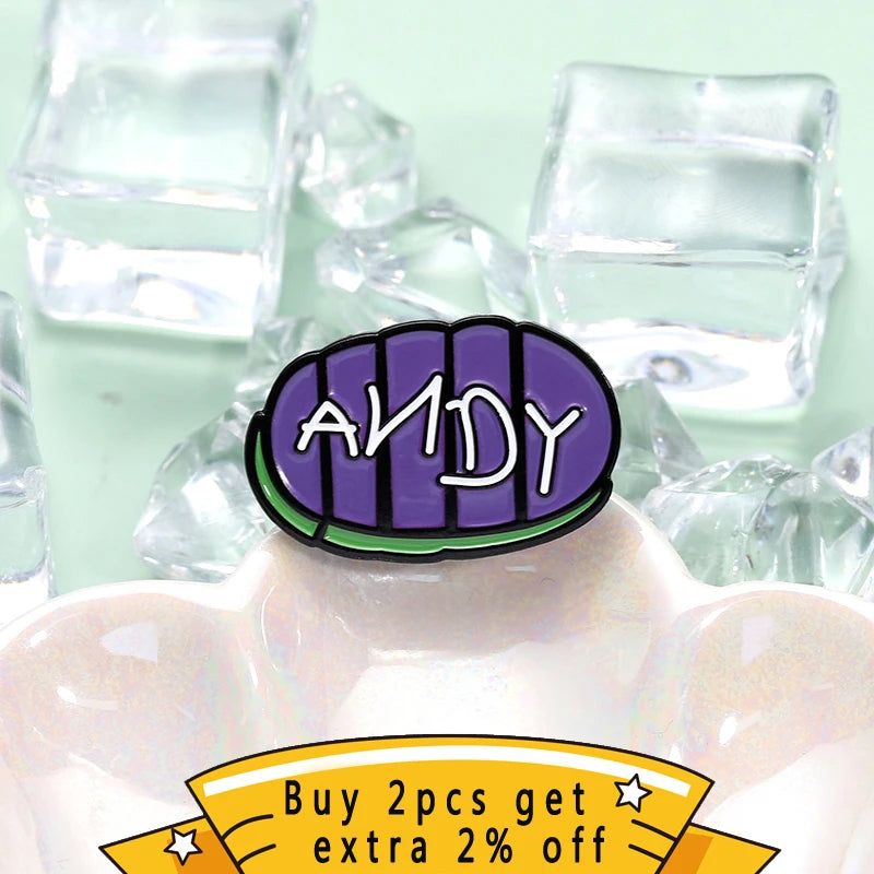 دبوس طية ياقة يحمل اسم ANDY بخط بنفسجي على خلفية من الإناء الوردي الفاتح.