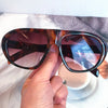 A hand holds a pair of vintage gradient leopard sunglasses with brown and amber lenses on a white surface.