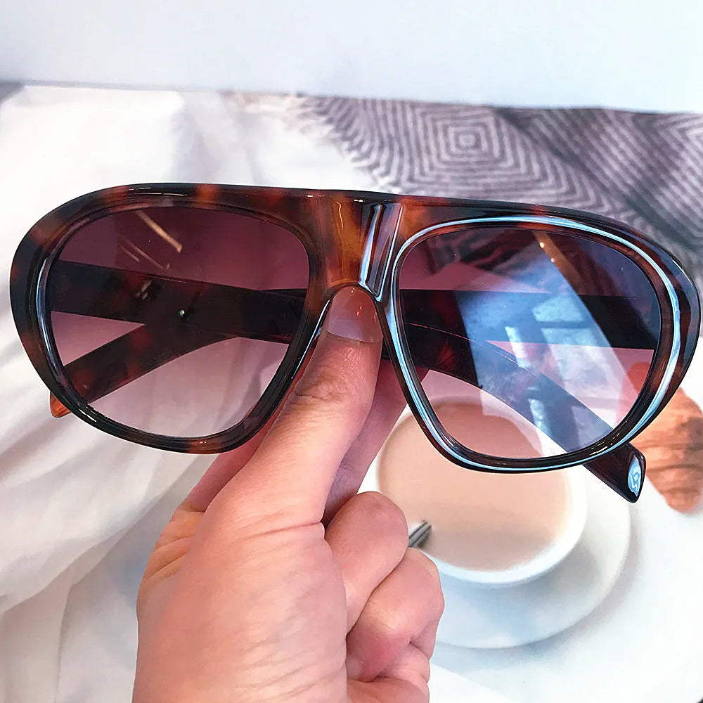 A hand holds a pair of vintage gradient leopard sunglasses with brown and amber lenses on a white surface.