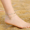 Gold plated initial letter anklet on a person's ankle resting on sand