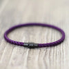 Purple thread charm bracelet with a black clasp resting on a light wooden surface.
