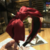 A deep red velvet wide bow headband is shown being held, with a photo on a wooden table in the background.