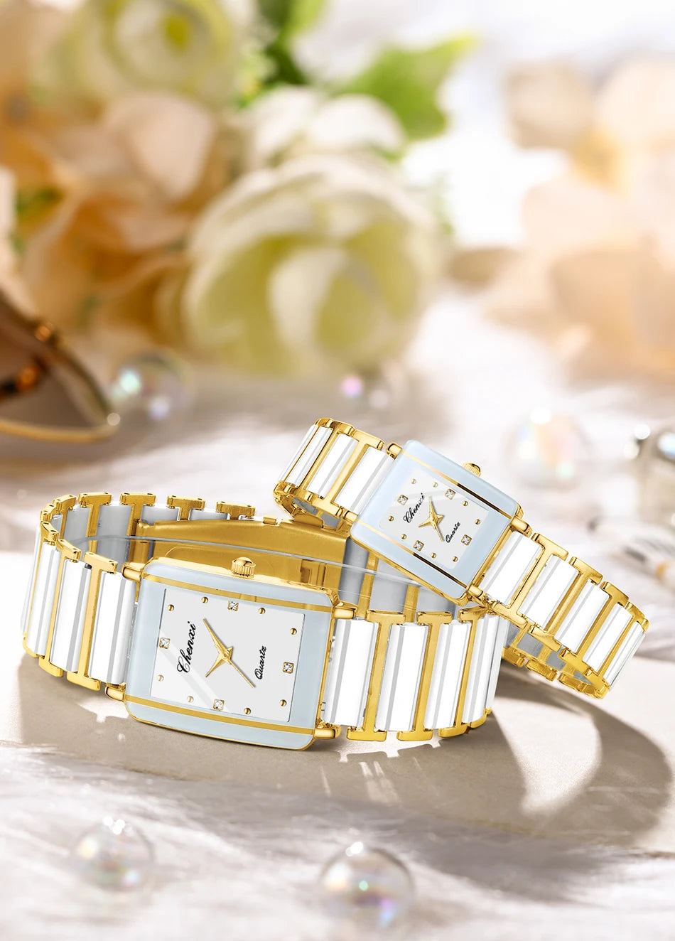 A pair of gold and white ceramic fashion wristwatches displayed on a light surface with soft floral background.