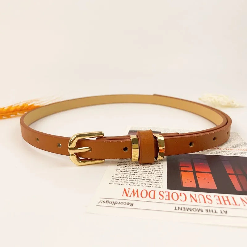A brown slim PU leather belt with a vintage gold-toned buckle lying on a piece of paper.