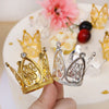 A hand holds a gold and silver mini crown cake topper against a blurred background of pink flowers and dark berries.