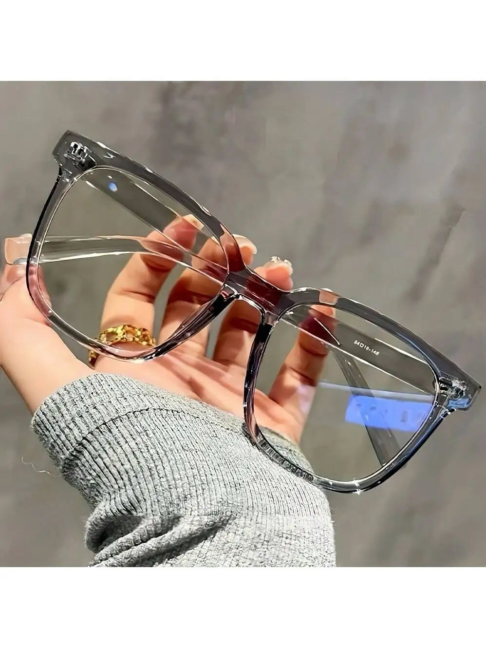 A hand holds a pair of clear square-shaped eyeglasses with thin pink frames against a gray background.