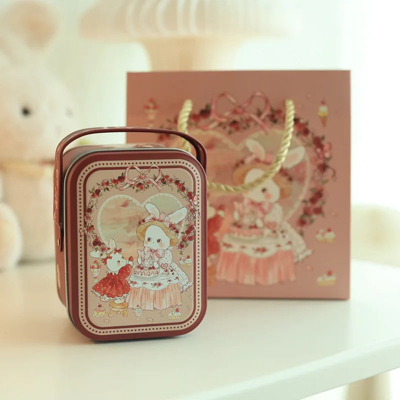 A small brown tinplate candy box with a carrying loop sits in front of a larger pink decorative box featuring a white rabbit 