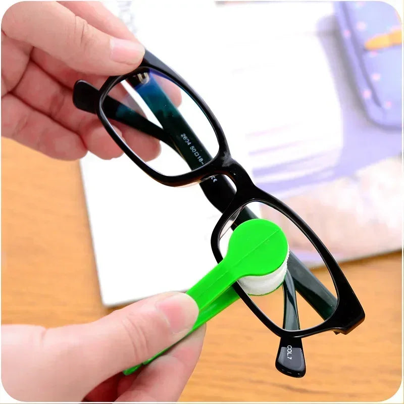 A hand holds a black frame eyeglasses cleaning kit with a green cleaning tool and white cleaning pad.